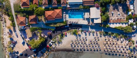 An aerial view of a coastal resort with multiple red-roofed buildings, a swimming pool, and a beach area with umbrellas.