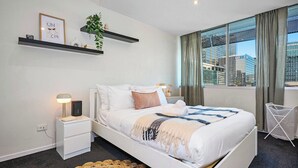A modern hotel room with a bed, bedside tables, a lamp, a plant, and a view of buildings through the window. - Steps away from South Cross - Easy 2B Urban Living (West Melbourne)