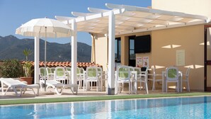 Area piscina con sedie a sdraio bianche, un pergolato con ombrelloni e vista sulle montagne.