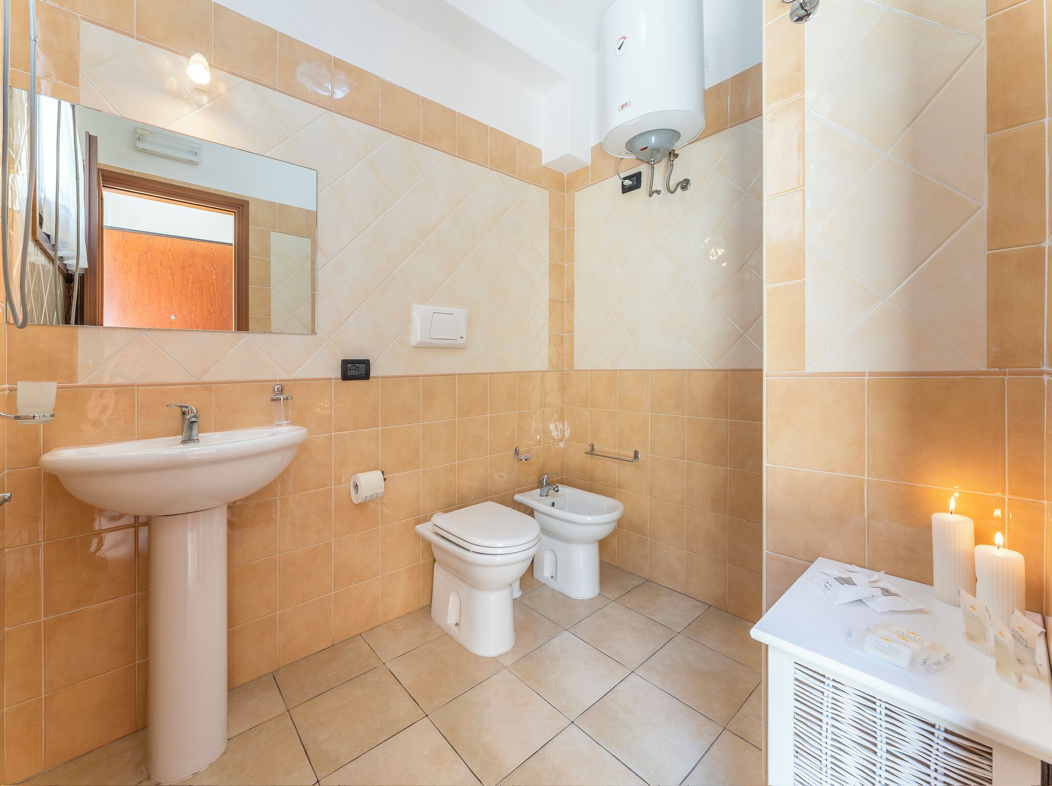 A bathroom with a sink, two toilets, a mirror, a heater, and a candle on a vanity.