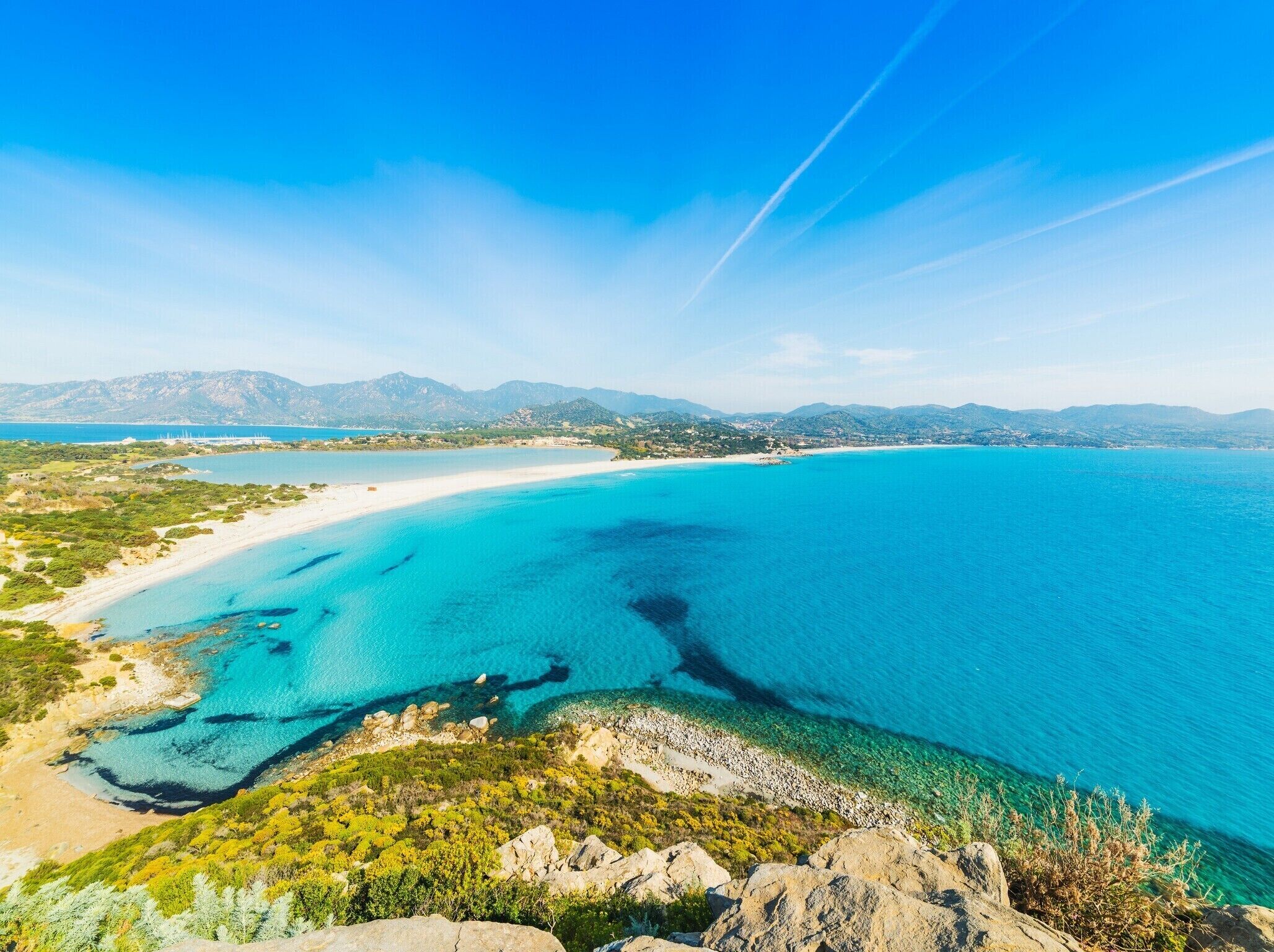 A coastal landscape with a clear blue sea, sandy beach, and distant mountains.