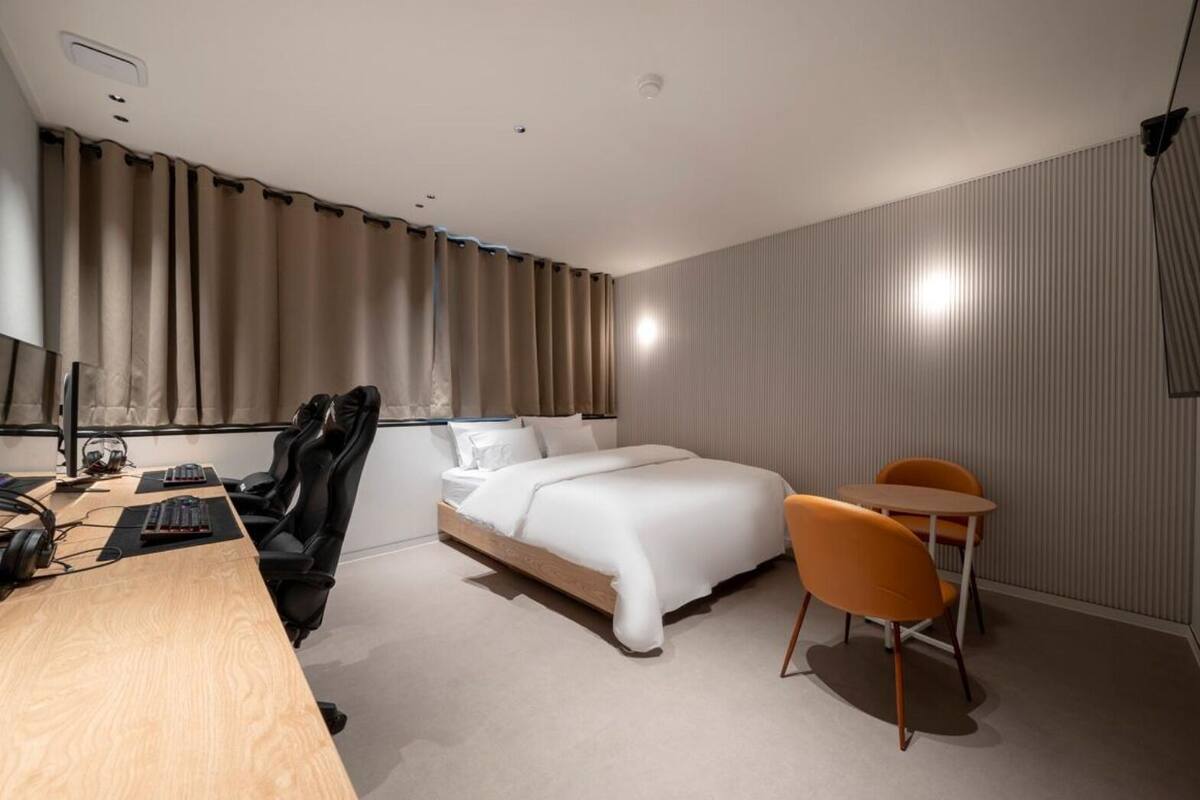 Una habitación de hotel moderna con un escritorio de madera, una silla de juego negra, una cama con ropa de cama blanca y una pequeña mesa naranja.