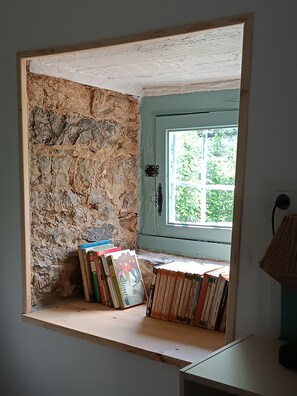 A room with a stone wall, a wooden shelf with books, and a window with a view of greenery. - Apartment for four people in nature (Grado)