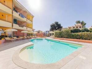 Une piscine entourée de chaises longues et de parasols, elle-même bordée d’un bâtiment doté de balcons, le tout sous un ciel dégagé.