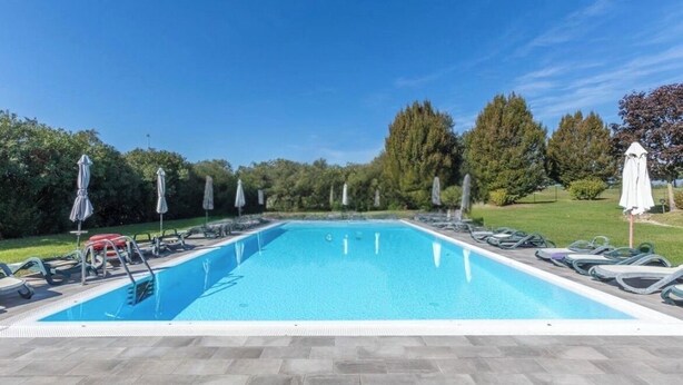 Una piscina rettangolare con bordo piastrellato, circondata da lettini e ombrelloni, con sullo sfondo alberi e un cielo sereno.
