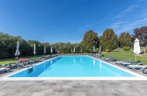 Une piscine rectangulaire, bordée d’une terrasse carrelée, entourée de chaises longues et de parasols, le tout se fondant dans un décor d’arbres et d’un ciel dégagé.