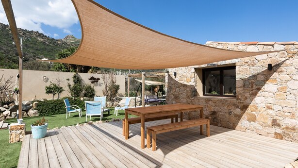 Una terraza de madera con una mesa y sillas, una vela de sombra, y un edificio de piedra con una ventana.