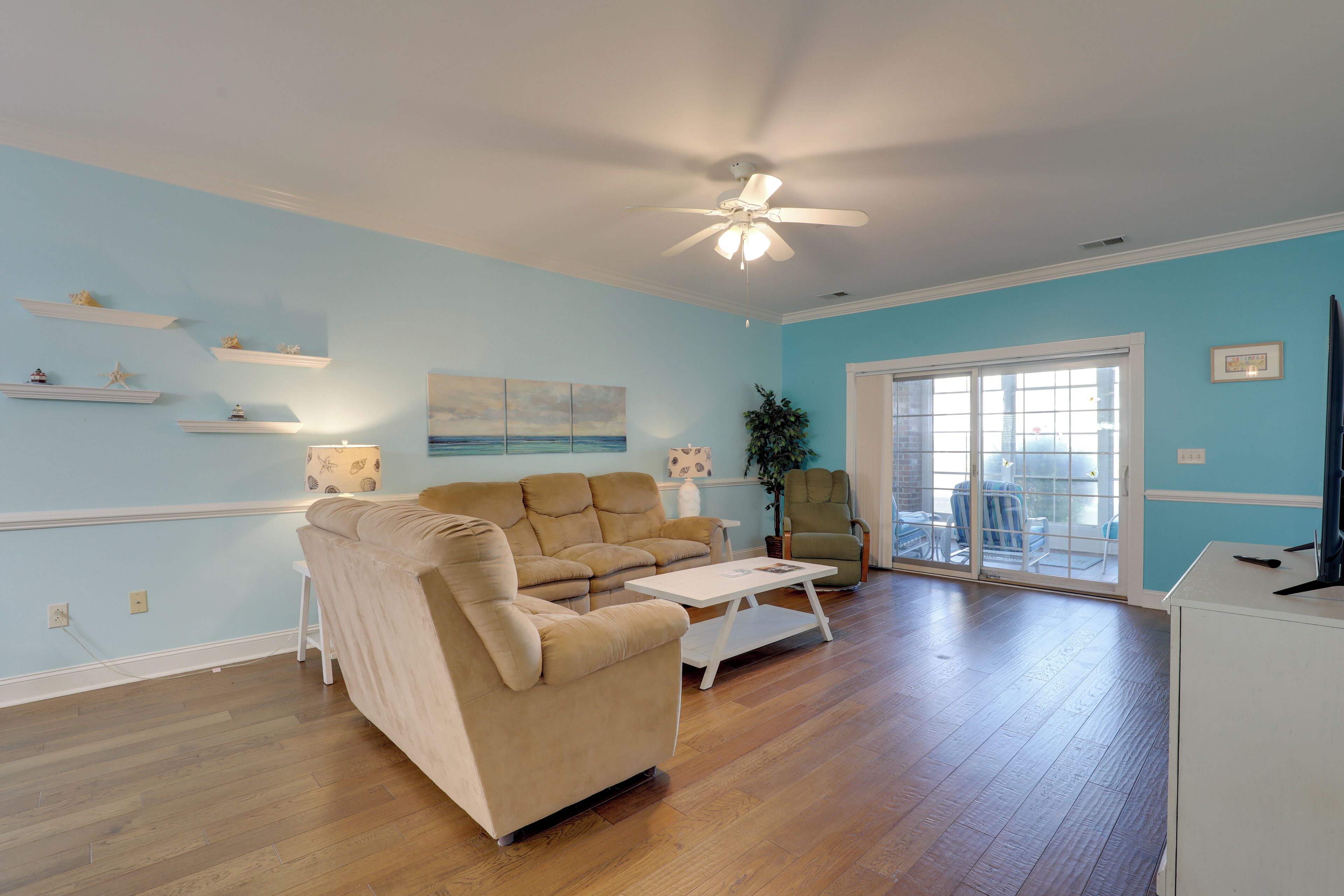 A living room with a beige sofa, a white coffee table, wooden flooring, a ceiling fan, and a sliding glass door leading to an outdoor area.
