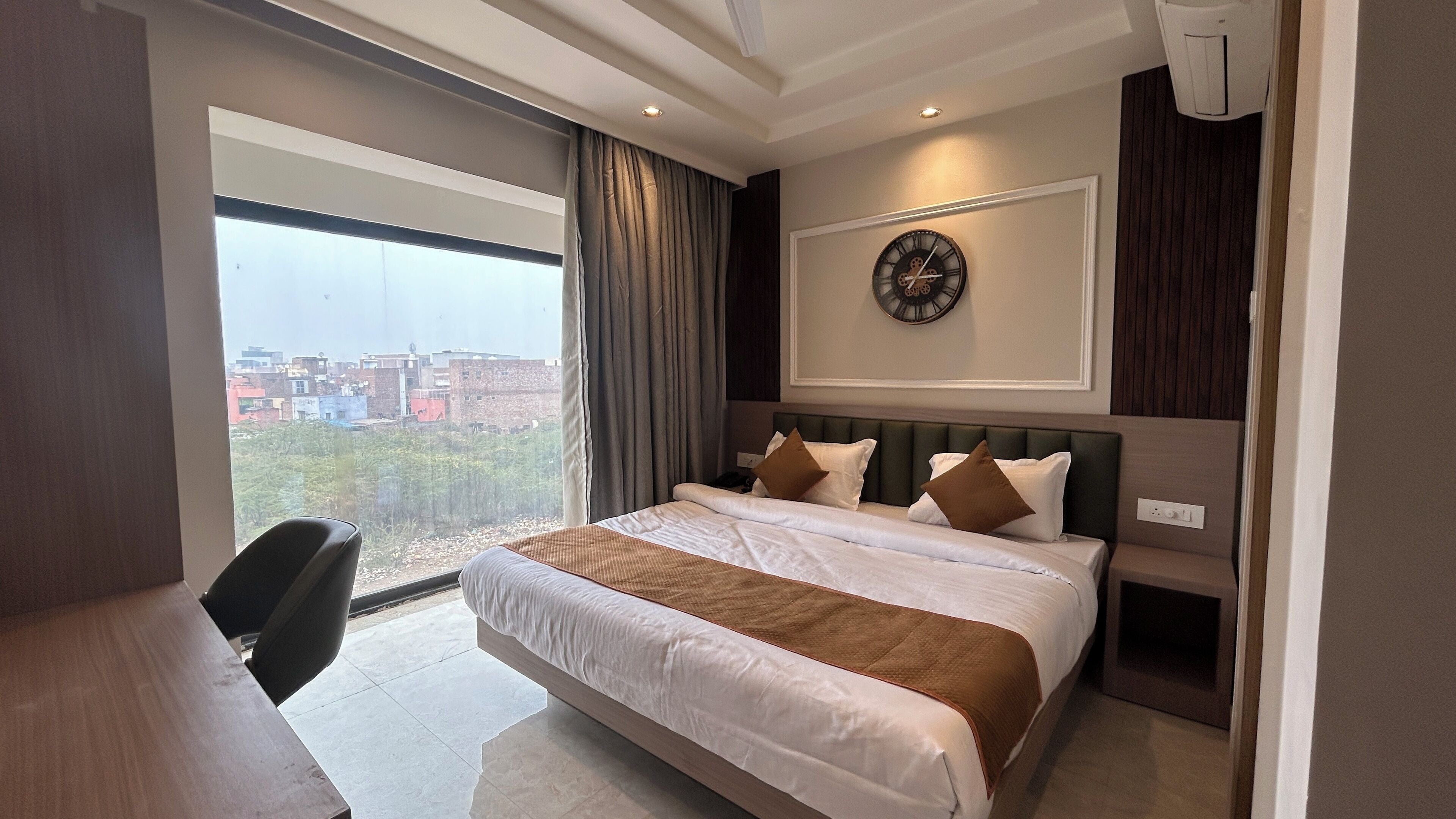 A hotel room with a large bed, a desk, a view of the city, and a clock on the wall.