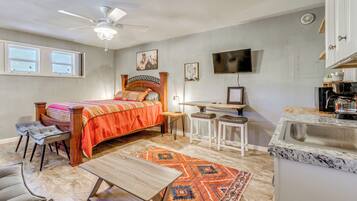 A bedroom with a bed, a desk, a chair, a small table, a TV, and a kitchen area with a sink.