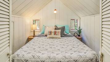 A cozy attic bedroom with a sloped ceiling, a bed with patterned bedding, two bedside tables with lamps, and a window with a view.