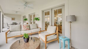 A living room with a sofa, armchair, and a small table with a plant. There are ceiling fans, a lamp, and a view of the kitchen through glass doors. - Beautiful Garden Villa With Golf Cart Option (Wildwood)