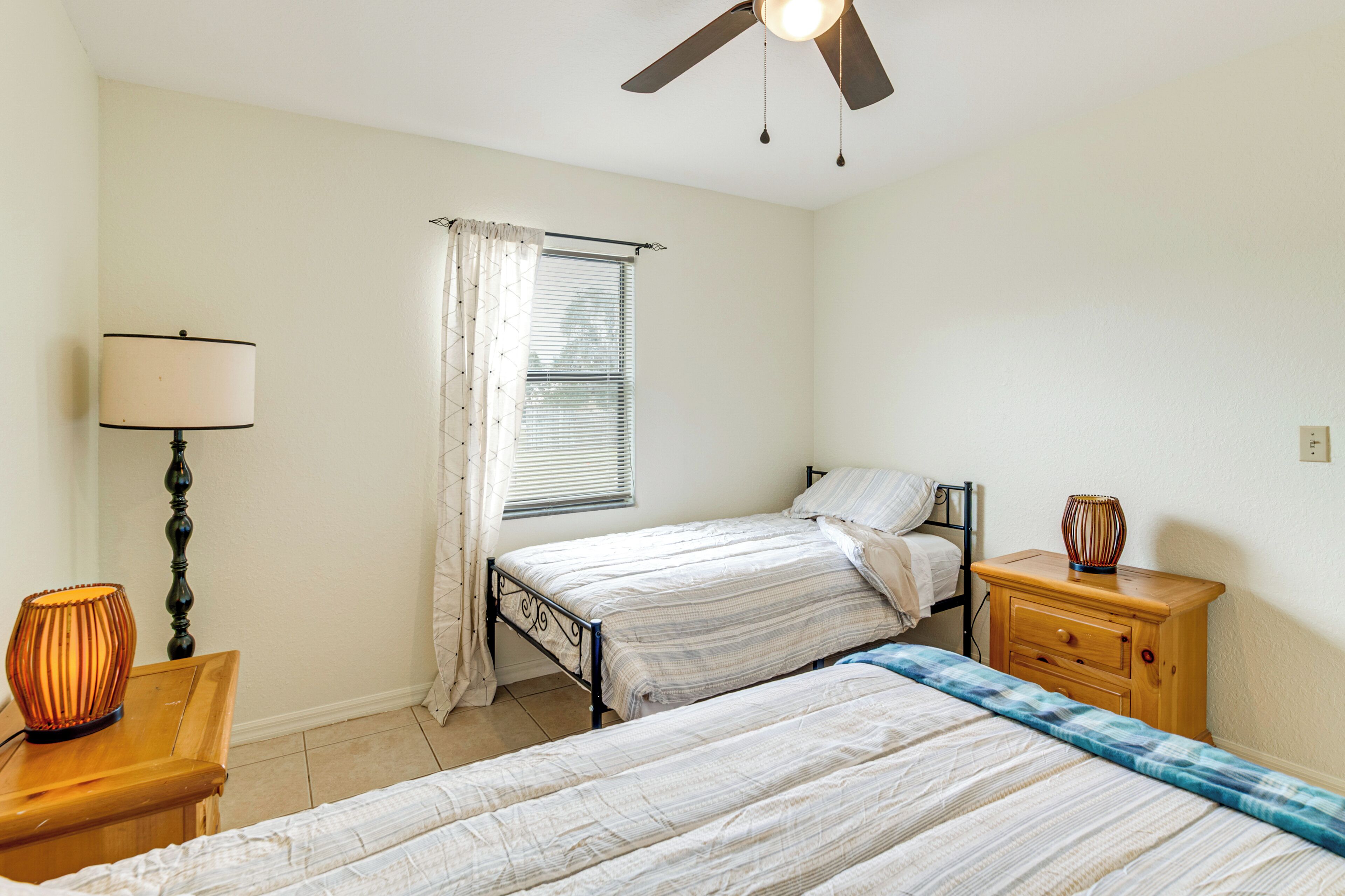 A bedroom with two beds, a ceiling fan, a bedside table with a lamp, and a window with curtains.