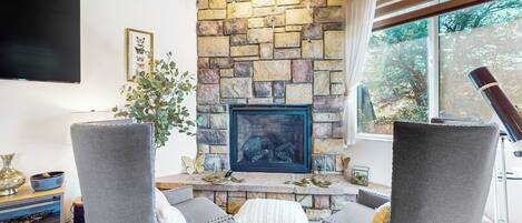 A living room with a stone fireplace, grey upholstered chairs, a flat-screen TV, and a window with a view of trees.