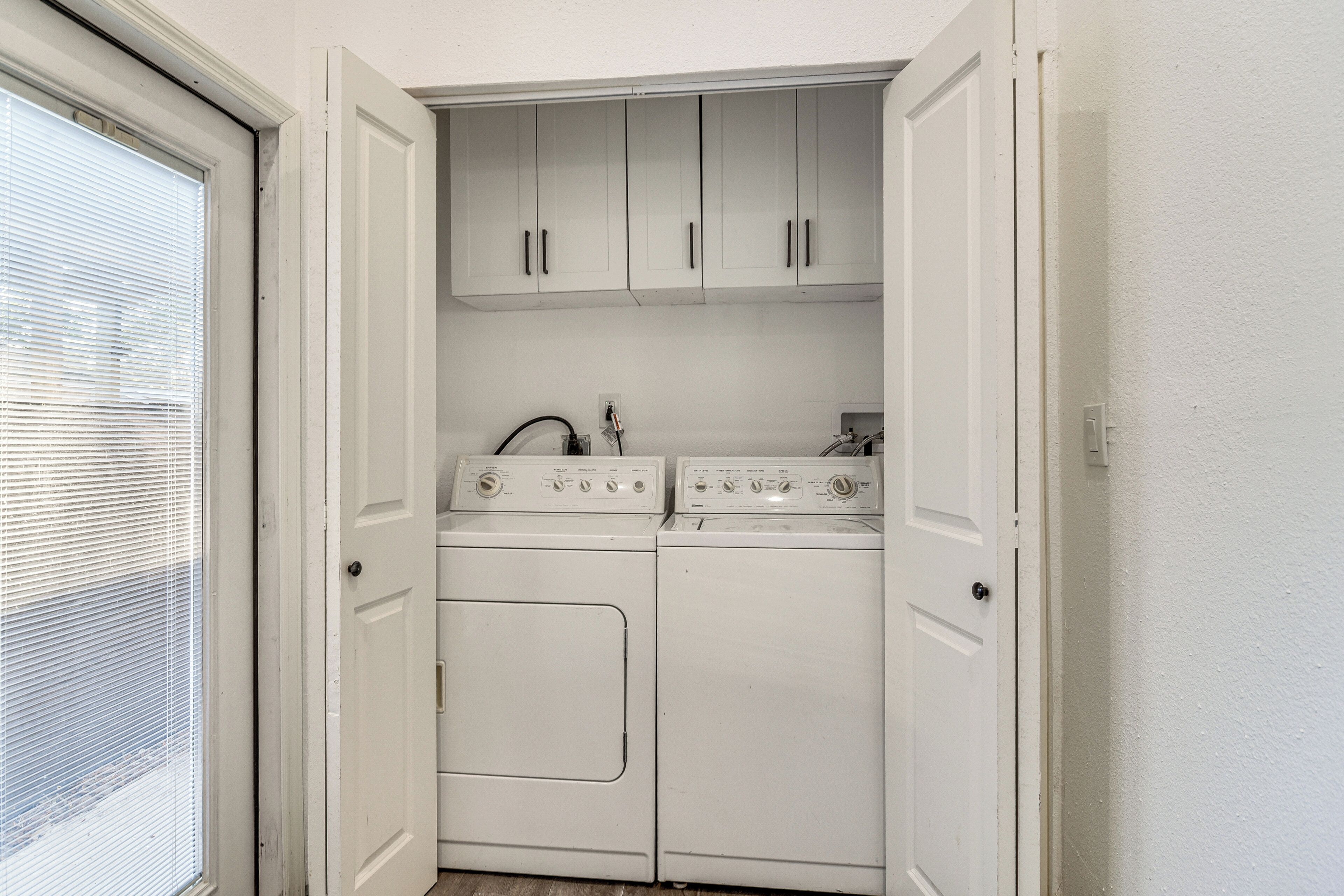 Une buanderie équipée d’un lave-linge et d’un sèche-linge, avec des placards blancs au-dessus, et une porte donnant sur une fenêtre munie de stores.