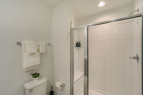 Un baño moderno con ducha envuelta en vidrio, azulejos blancos, inodoro, lavamanos y una pequeña planta en maceta.