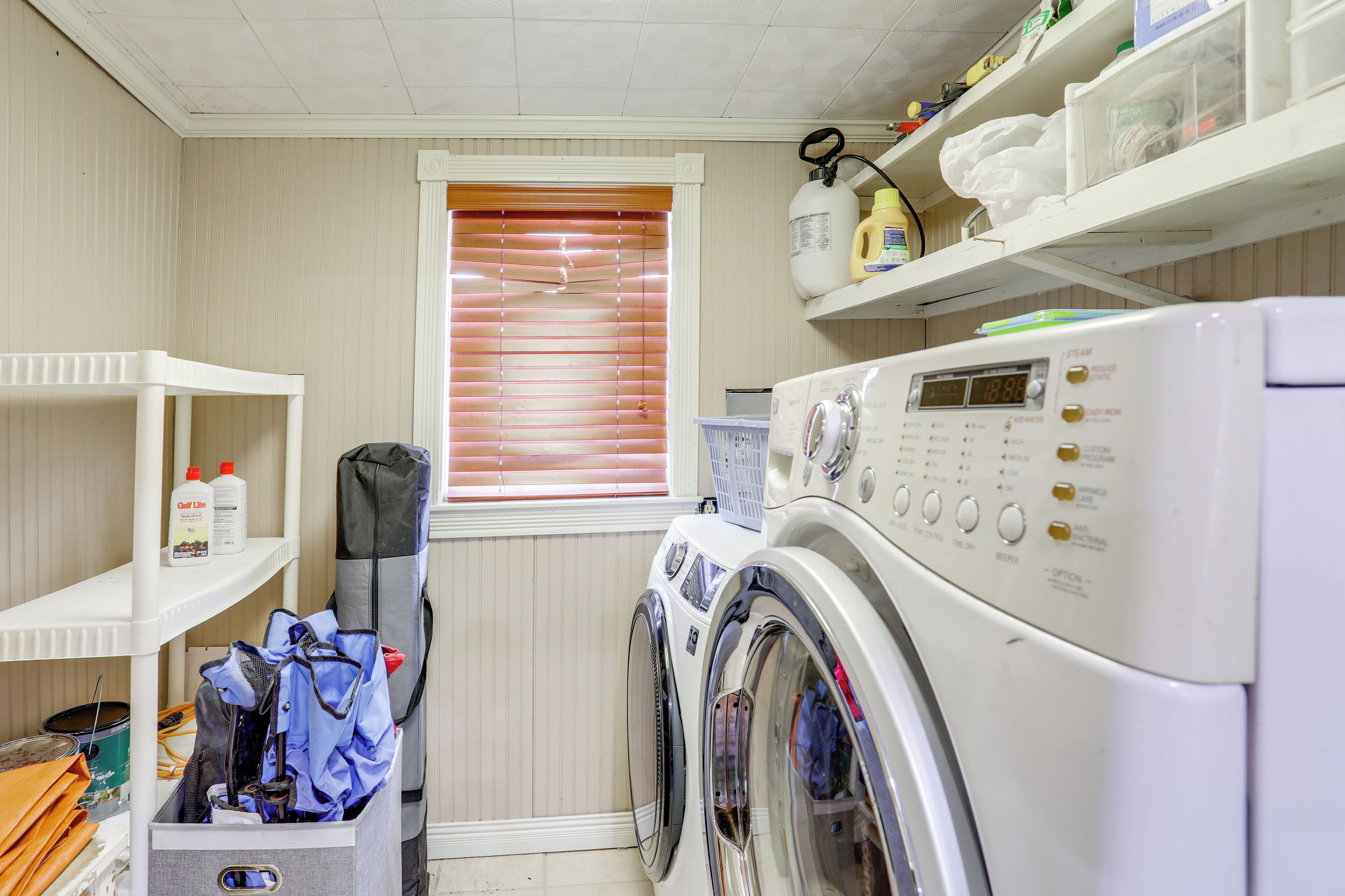 A laundry room with a front-loading washing machine, shelves with cleaning supplies, and a window with blinds.