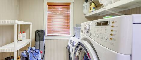 A laundry room with a front-loading washing machine, shelves with cleaning supplies, and a window with blinds.