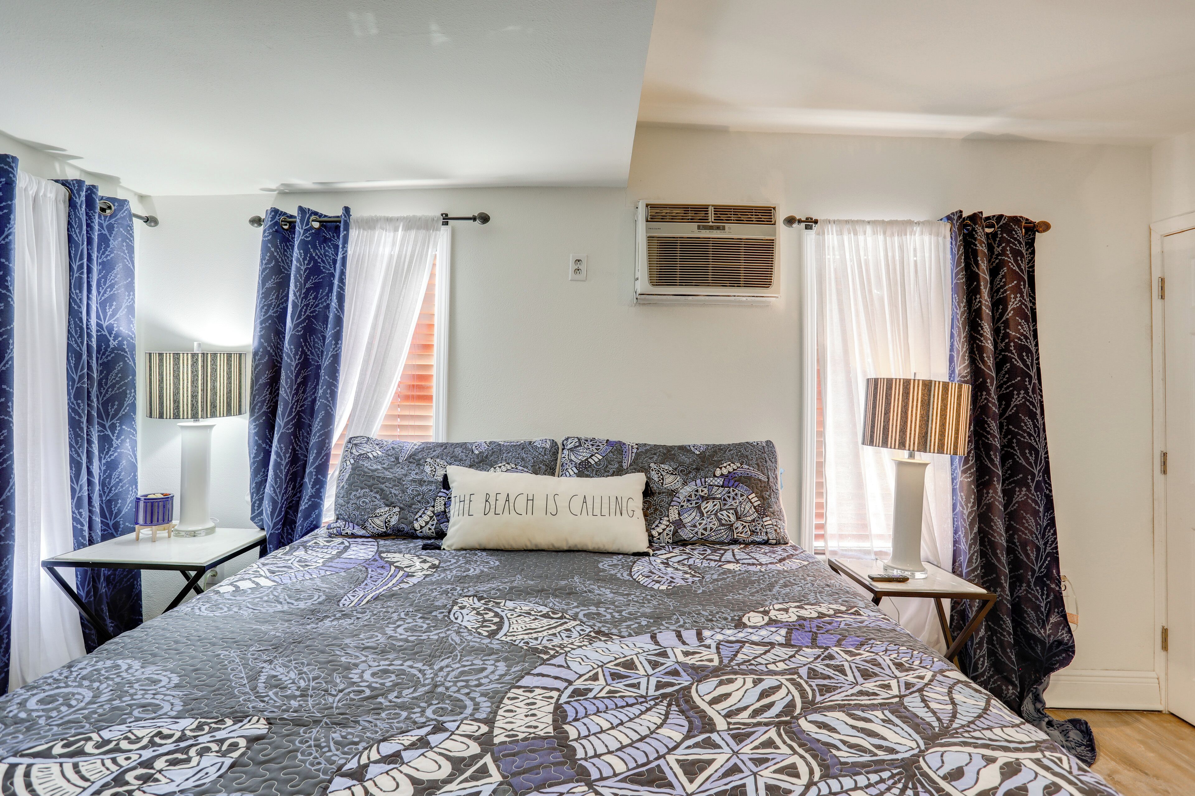 Un dormitorio con una colcha estampada, un almohadón con texto, una mesita de noche con lámpara y cortinas.
