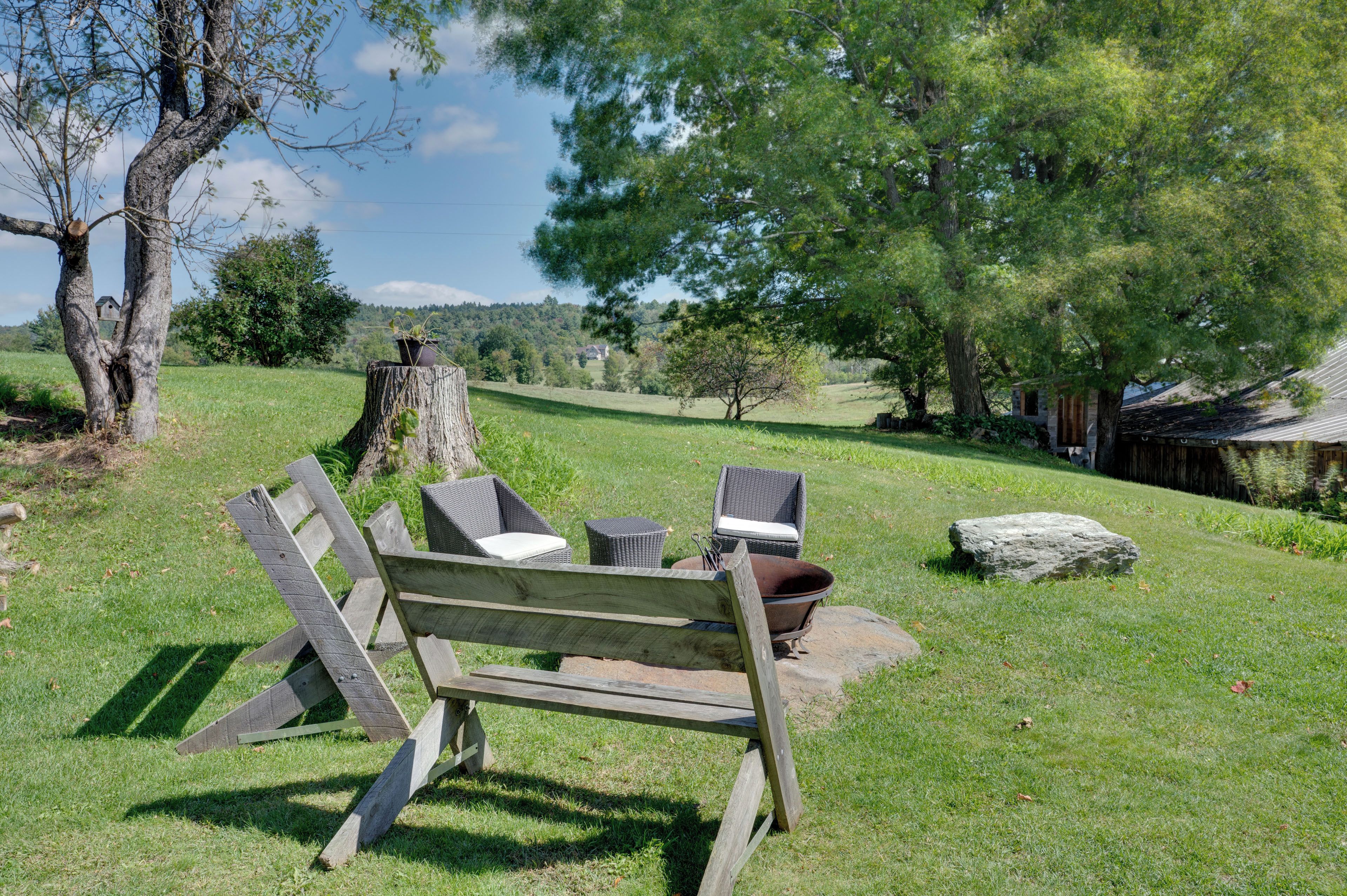 Un banc en bois, une chaise en osier et une petite table, installés sur un champ herbeux avec vue sur des arbres et un bâtiment situé au loin.