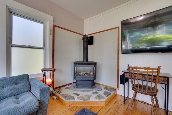A cozy living room with a wood-burning stove, a comfortable sofa, a wooden chair, and a flat-screen TV mounted on the wall. - Green Mountain Views! Lowell Getaway Near Skiing (Lowell)
