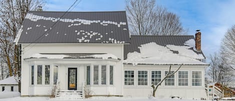 A two-story white house with a snow-covered roof and chimney, surrounded by bare trees.