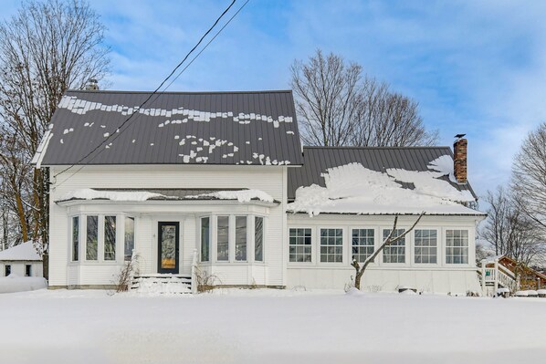 A two-story white house with a snow-covered roof and chimney, surrounded by bare trees. - Green Mountain Views! Lowell Getaway Near Skiing (Lowell)