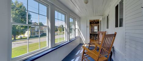 A covered porch with large windows, wooden rocking chairs, and a view of a street and houses.