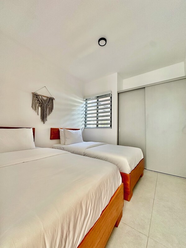 A modern bedroom with two beds, a wall-mounted macrame piece, and a window with blinds.