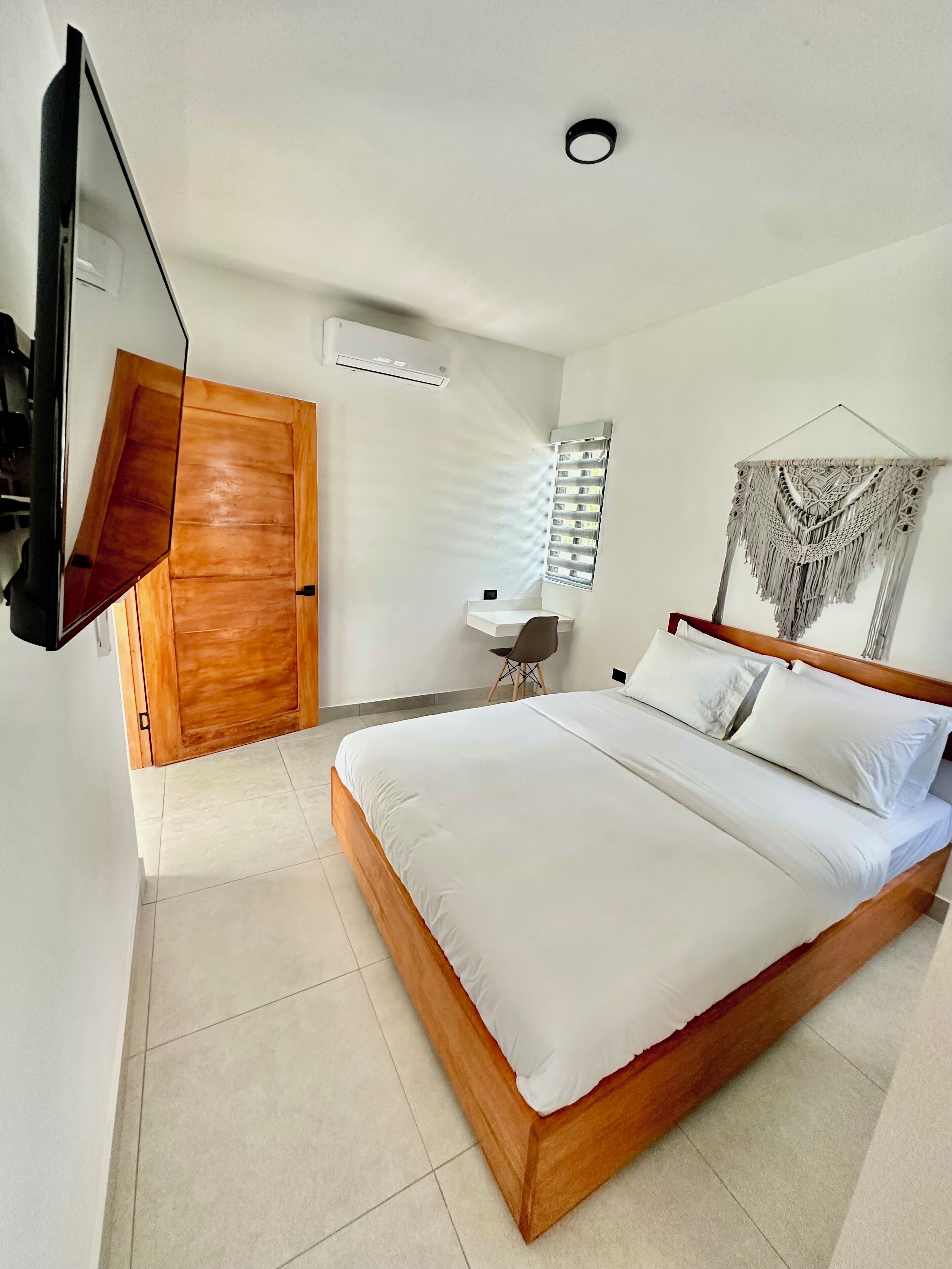 A modern bedroom with a wooden bed, a desk, and a macrame wall hanging.
