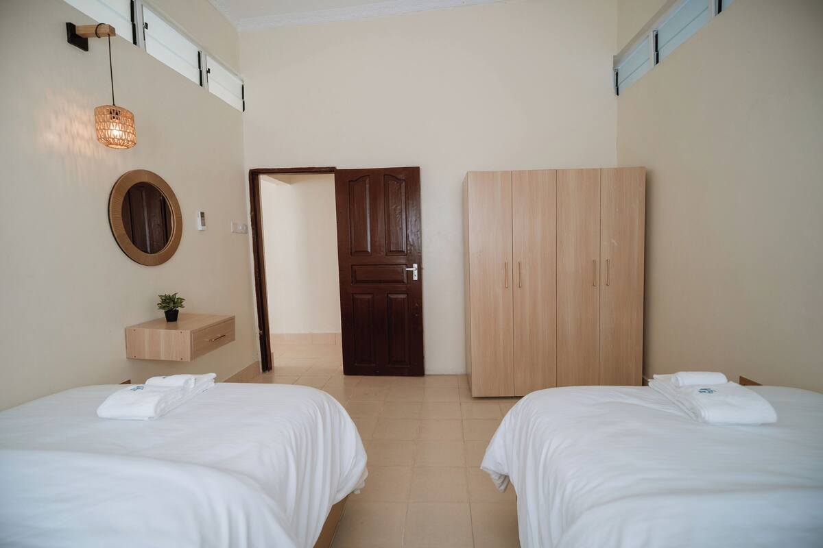 A hotel room with two beds, a wooden wardrobe, a wooden shelf with a plant, and a door leading to another room.