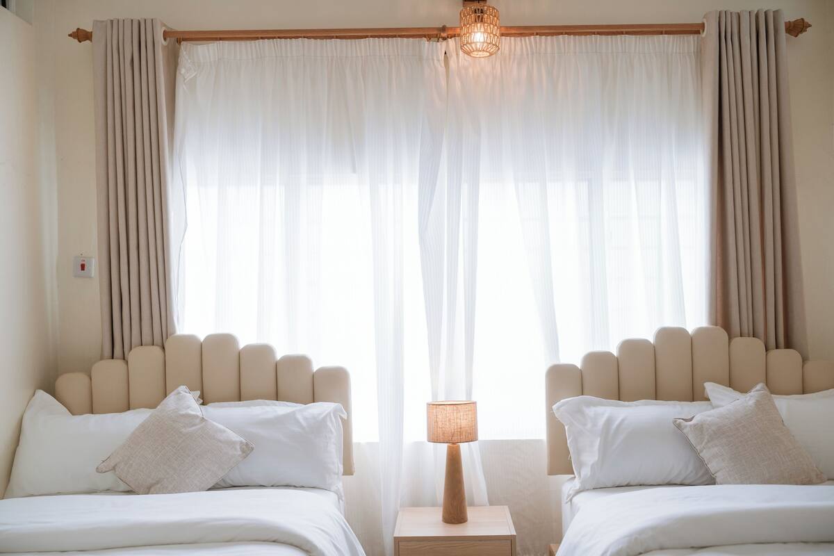 A hotel room with two beds, a bedside table with a lamp, and sheer curtains.