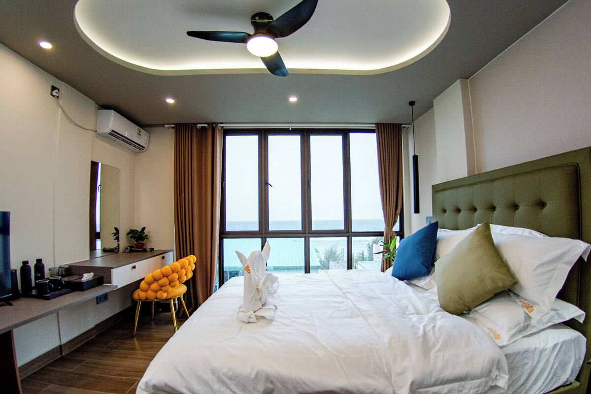 A hotel room with a bed, a desk, a chair, and a view of the sea through large windows.