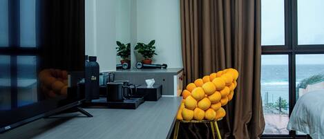 A hotel room with a TV, remote control, a desk with a plant, a chair, and a view of the sea.