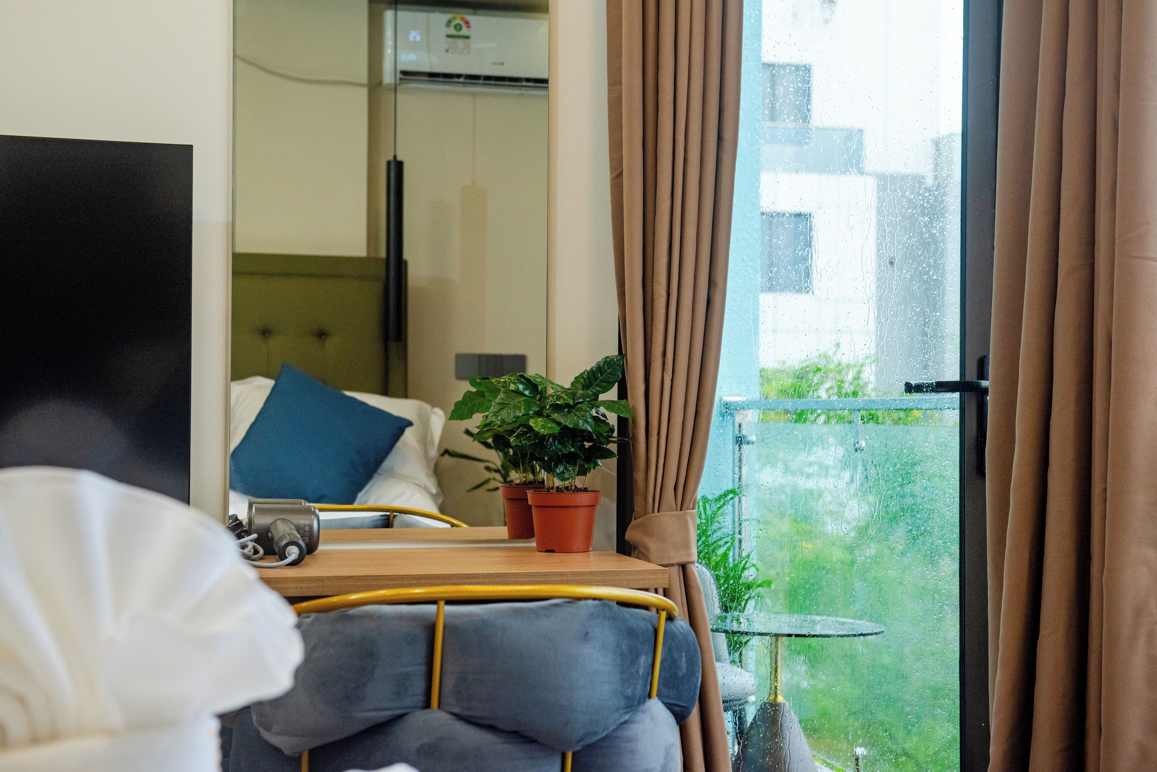 Habitación de hotel con cama, mesita de noche con planta, una pequeña mesa con lámpara y vistas al exterior a través de una ventana.