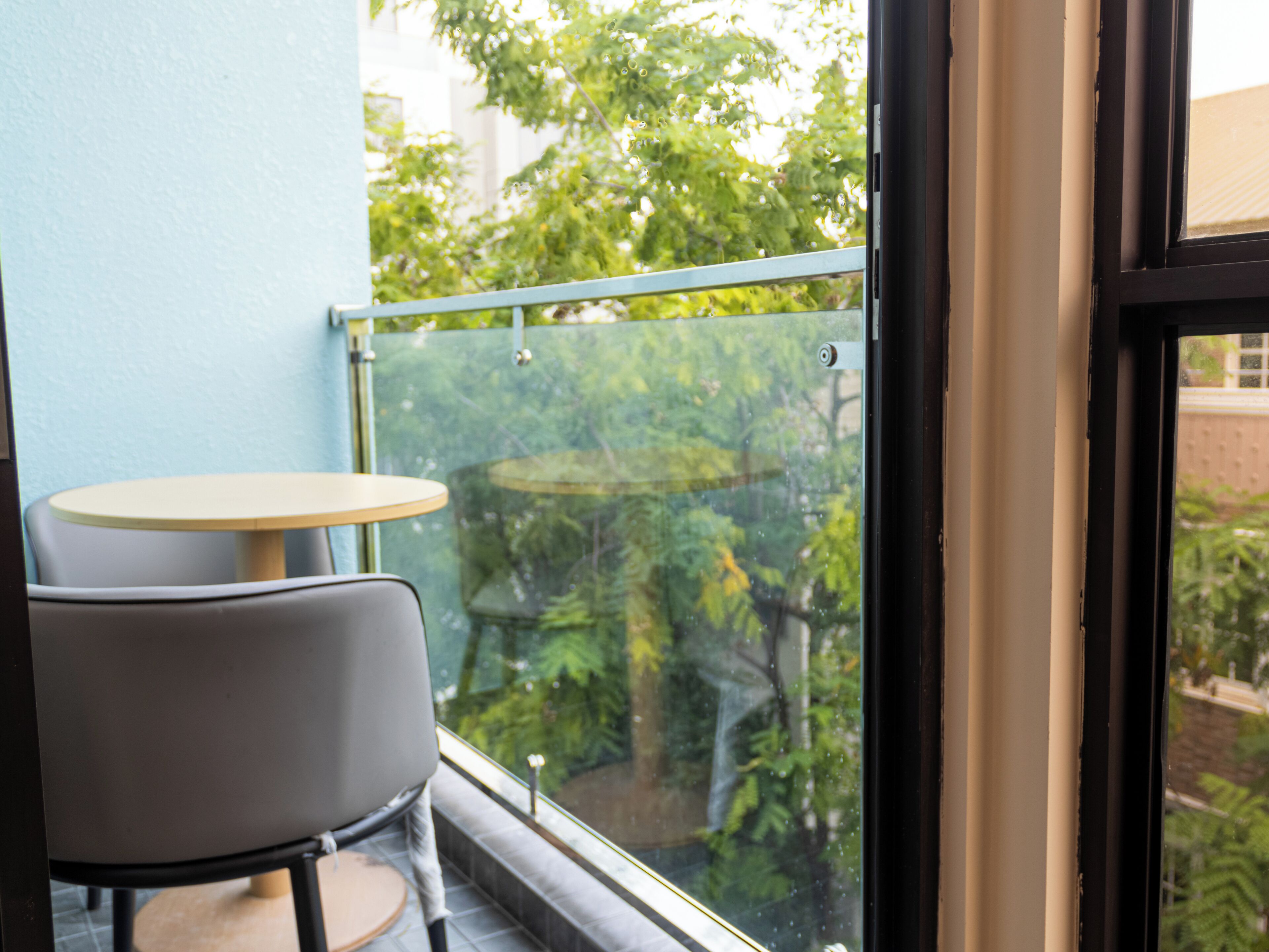 Un balcón con una mesa, una silla y barandilla de vidrio, y vistas a la vegetación.