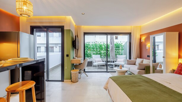 Ein modernes Hotelzimmer mit einer Küchenzeile, einem Bett, einer Sitzecke inklusive Sofa und Sesseln sowie einer großen Glasschiebetür, die auf einen Außenbalkon führt.