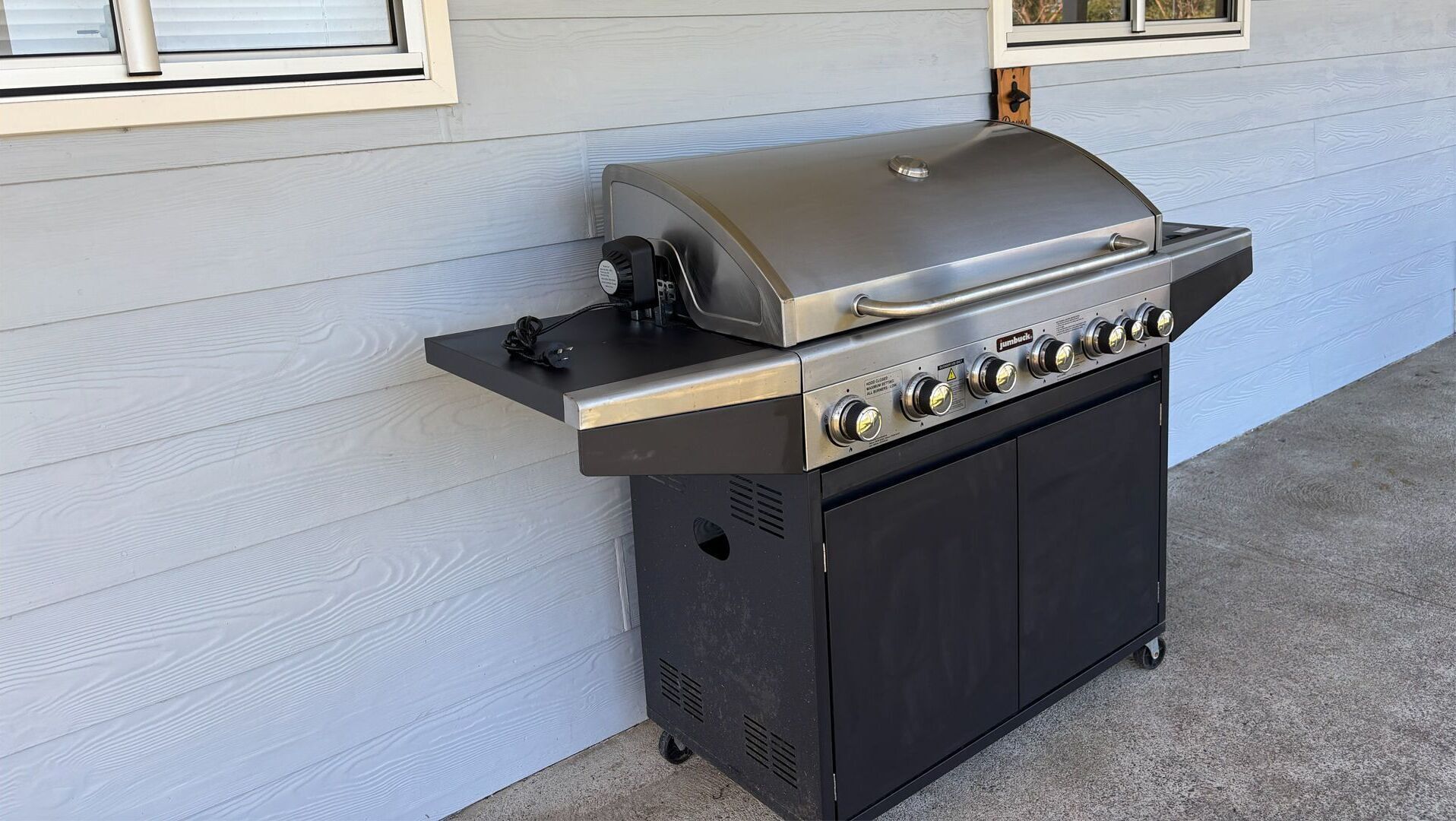 A portable gas grill with a control panel and a lid, placed against a house wall.