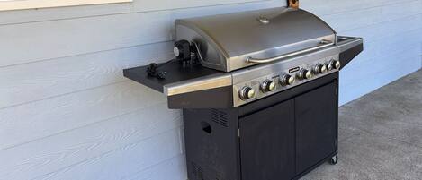 A portable gas grill with a control panel and a lid, placed against a house wall.