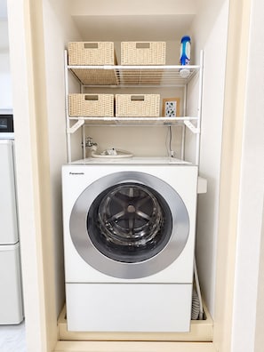 A compact washing machine with a front-loading design, placed in a small laundry room with a white shelf above it holding baskets and a blue bottle. - Easy Access to Shinjuku , Shibuya Area , 4 Guests , Studio (Shibuya honmachi)