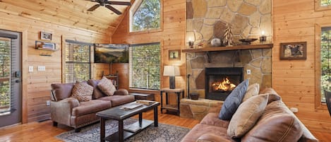 Un acogedor interior de cabaña de troncos con chimenea de piedra, muebles de madera y ventanas grandes.