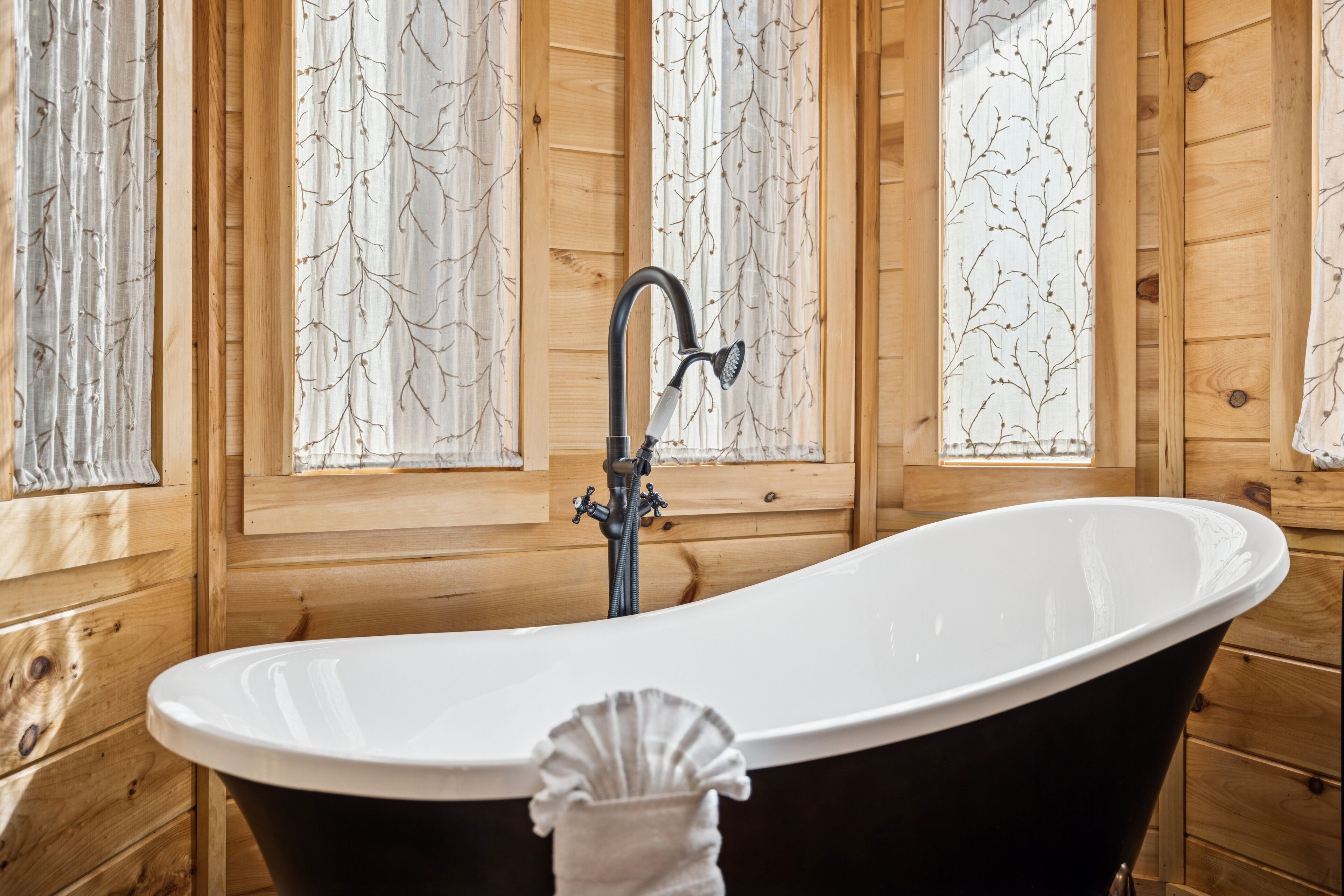 Baño de cabaña de madera con bañera independiente, paredes de madera y ventanas con vidrio esmerilado.