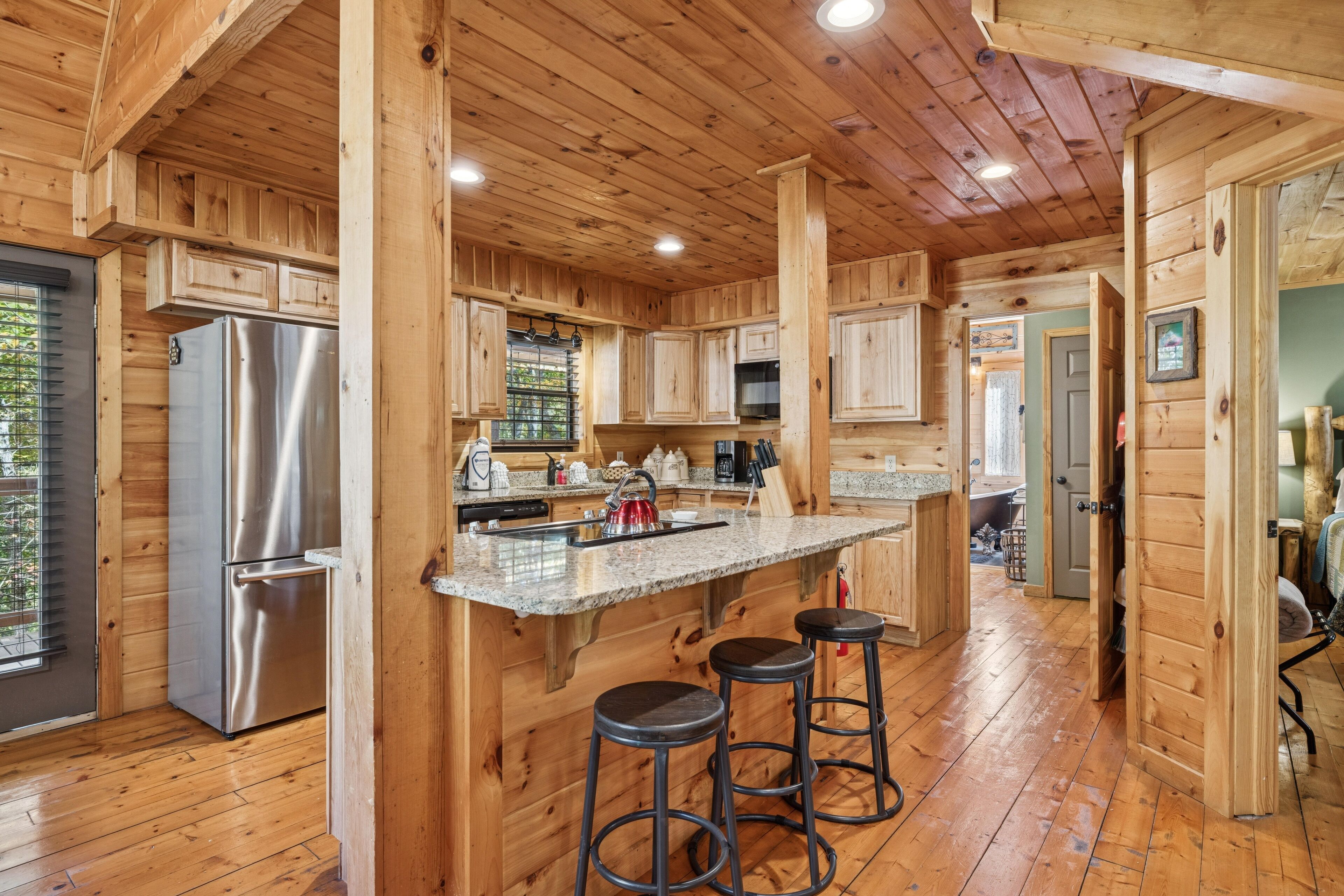 A wooden cabin kitchen with a stainless steel refrigerator, granite countertop, and wooden stools.
