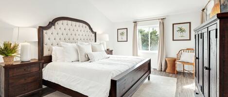 Ein Schlafzimmer mit einem großen Bett, Nachttischen aus Holz, einer Kommode und einem Fenster mit Vorhängen.