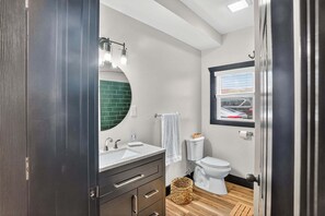 Une salle de bain moderne avec une porte en bois foncé, un lavabo blanc avec un miroir, un mur recouvert de carreaux verts, des toilettes blanches et une fenêtre donnant sur une voiture.