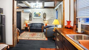 A cozy living area with a black leather sofa, a blue armchair, and a wooden coffee table. A patterned rug covers the floor, and a painting hangs on the wall.