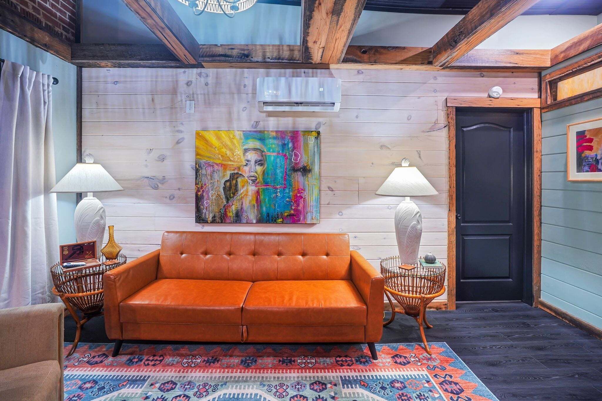 A room with a leather sofa, two side tables with lamps, a colorful abstract painting, and a patterned rug.