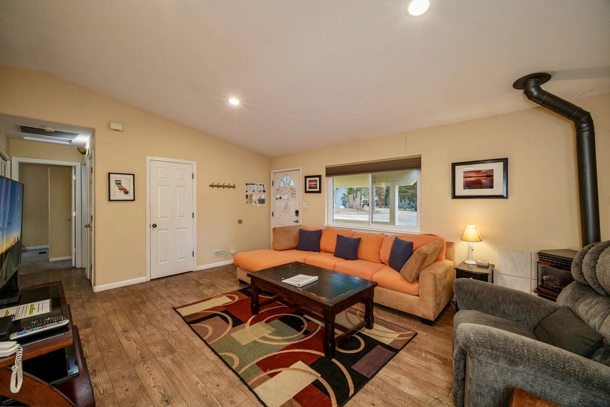 A living room with a beige sofa, a coffee table, and a gray armchair. There is a window with a view of the outside, a door, and a wall-mounted lamp.