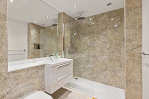 A modern bathroom with a glass-enclosed shower, a white vanity with a mirror, and marble walls. - The Ford - One Bed Apt (Chelmsford)