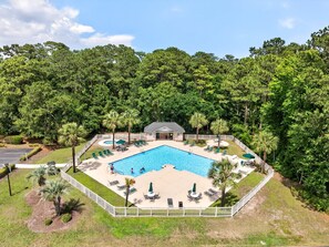 Piscina rectangular con tumbonas y palmeras, rodeada por una cerca blanca y un bosque denso.